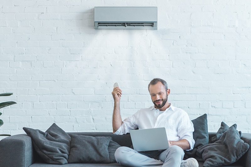 4 Amazing Benefits of Ductless Units. Man in his home, sitting on his couch on his laptop, turning on a ductless unit above him.